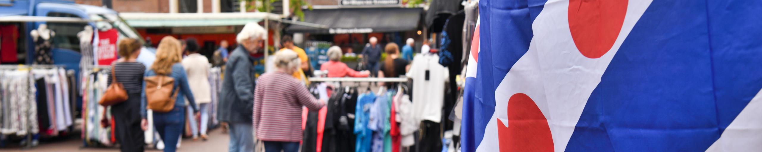 op de voorgrond een Friese vlag en op de achtergrond de markt in Leeuwarden met mensen die marktkraampjes bekijken